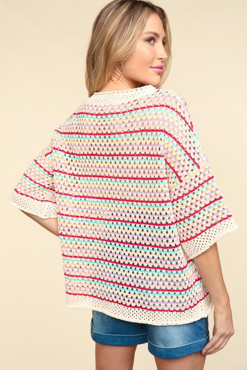 A woman stands smiling against a beige background, wearing the Haptics Striped Crochet Drop Shoulder Knit Top paired with denim shorts.