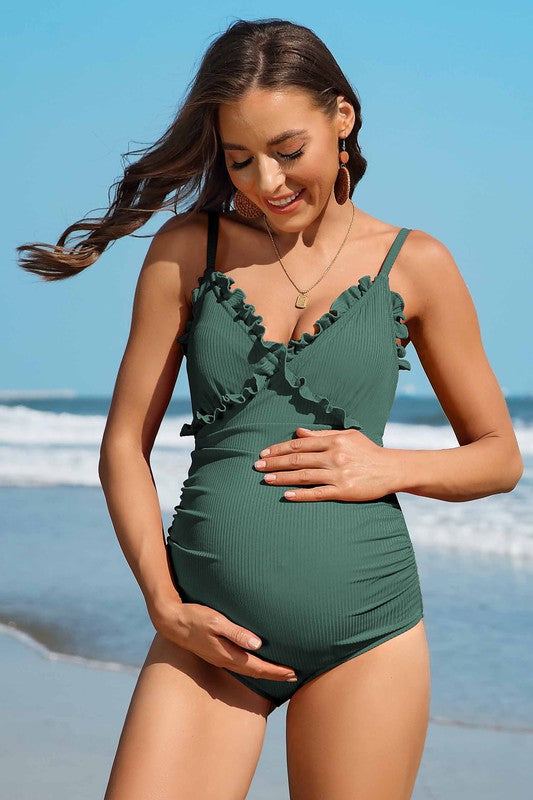 A woman in a Ruch Floral Maternity Swimsuit Pregnancy Swimwear stands on the beach, smiling and holding her pregnant belly. The ocean is visible in the background.