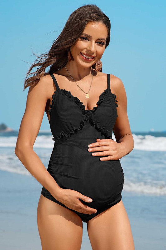 A woman in a Ruch Floral Maternity Swimsuit Pregnancy Swimwear stands on the beach, smiling and holding her pregnant belly. The ocean is visible in the background.