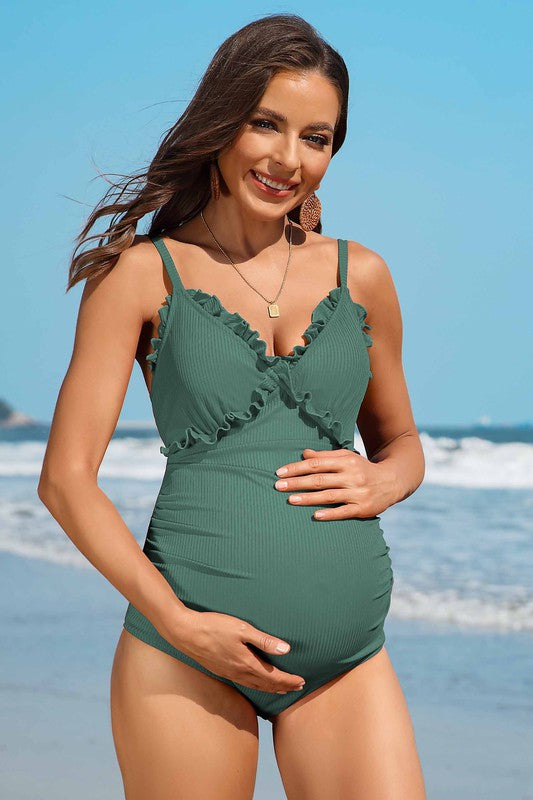 A woman in a Ruch Floral Maternity Swimsuit Pregnancy Swimwear stands on the beach, smiling and holding her pregnant belly. The ocean is visible in the background.