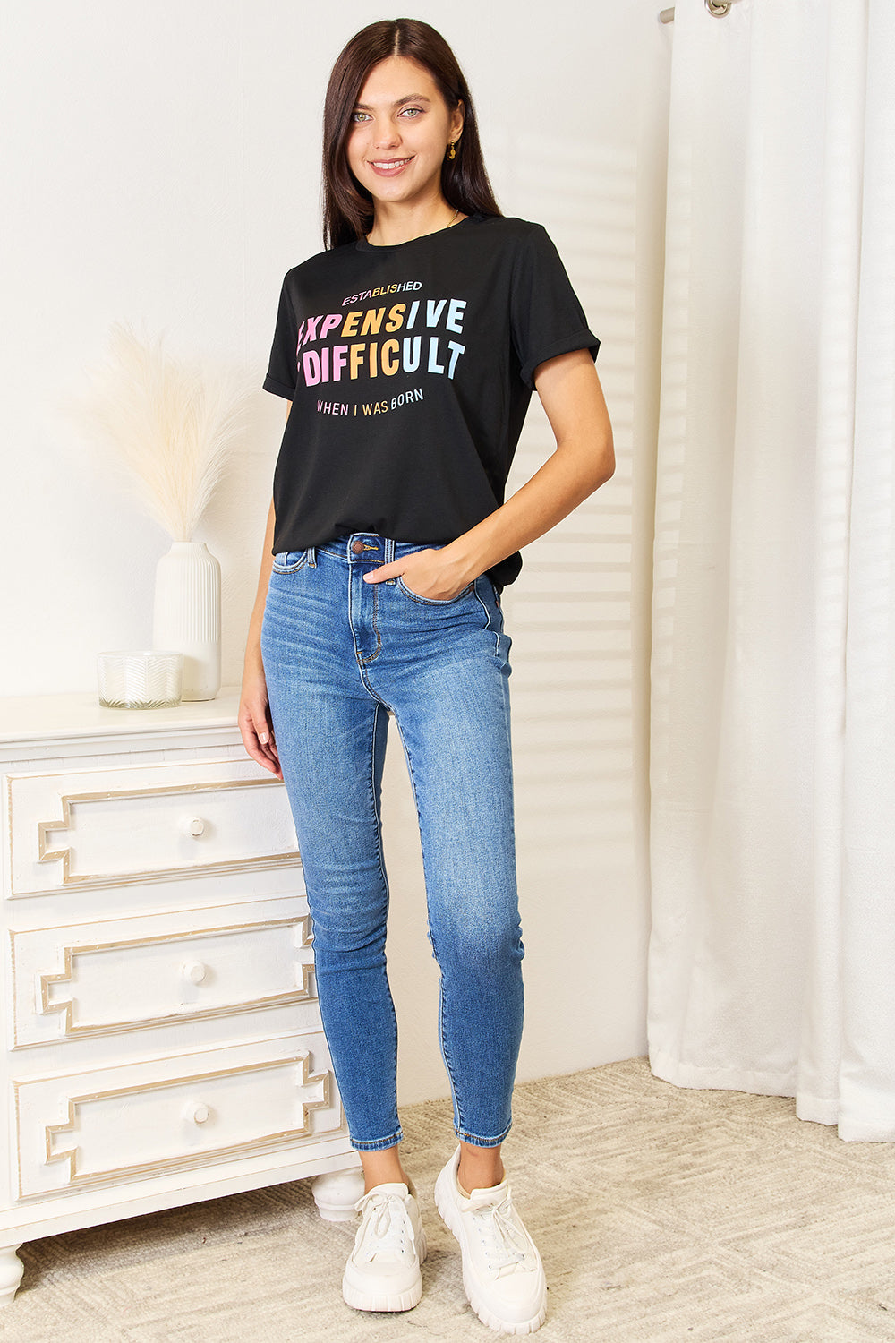 A woman with long braided hair, wearing the Simply Love Slogan Graphic Cuffed Sleeve T-Shirt that says "Established Expensive & Difficult When I Was Born," stands with one hand in her pocket. The trendy cuffed sleeves of her shirt add an extra touch of style.