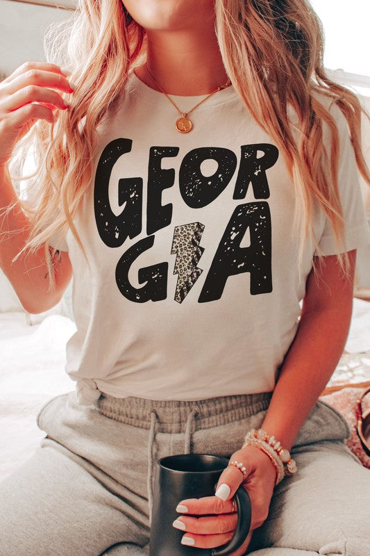 A person wearing a white GEORGIA LEOPARD LIGHTNING GRAPHIC TEE, featuring the word "GEORGIA" and a lightning bolt design, is seated while holding a black mug. Their long, wavy blonde hair cascades down their shoulders, highlighting the unisex sizing of this stylish piece.