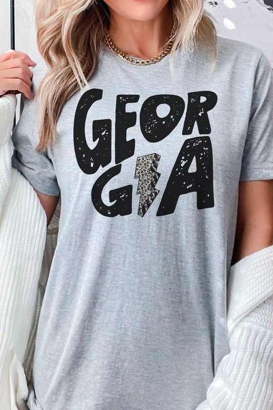 A person wearing a white GEORGIA LEOPARD LIGHTNING GRAPHIC TEE, featuring the word "GEORGIA" and a lightning bolt design, is seated while holding a black mug. Their long, wavy blonde hair cascades down their shoulders, highlighting the unisex sizing of this stylish piece.