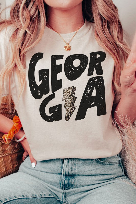 A person wearing a white GEORGIA LEOPARD LIGHTNING GRAPHIC TEE, featuring the word "GEORGIA" and a lightning bolt design, is seated while holding a black mug. Their long, wavy blonde hair cascades down their shoulders, highlighting the unisex sizing of this stylish piece.