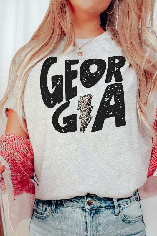 A person wearing a white GEORGIA LEOPARD LIGHTNING GRAPHIC TEE, featuring the word "GEORGIA" and a lightning bolt design, is seated while holding a black mug. Their long, wavy blonde hair cascades down their shoulders, highlighting the unisex sizing of this stylish piece.