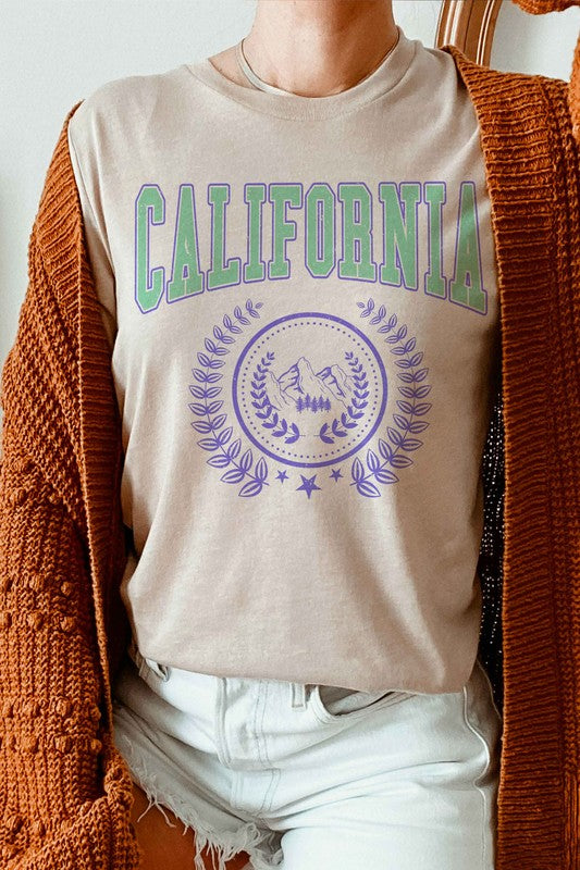 Person wearing a CALIFORNIA GRAPHIC TEE, which features "California" written in green and purple letters along with a mountain and laurel wreath design, paired with white shorts. The T-shirt is unisex and available in plus sizes.