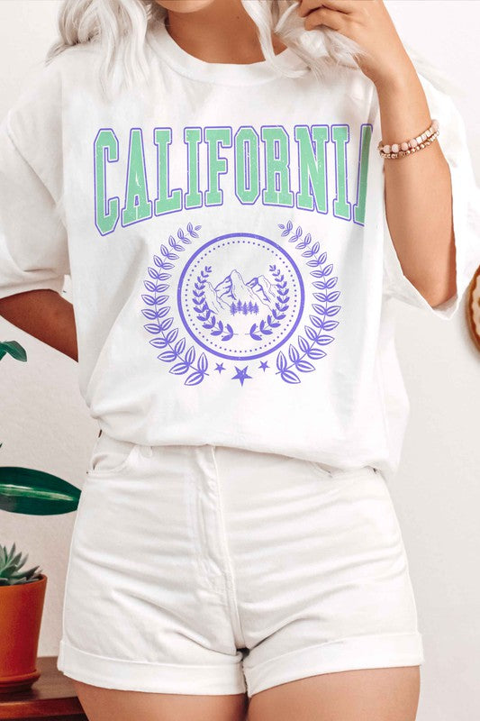 Person wearing a CALIFORNIA GRAPHIC TEE, which features "California" written in green and purple letters along with a mountain and laurel wreath design, paired with white shorts. The T-shirt is unisex and available in plus sizes.