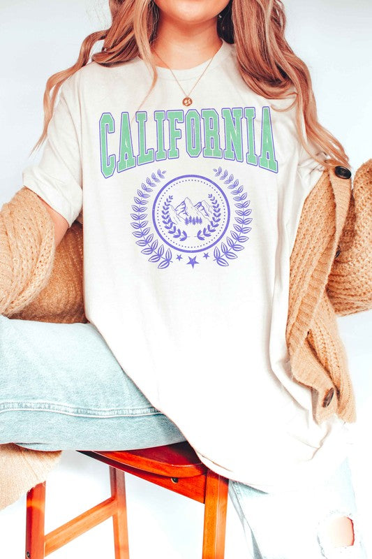 Person wearing a CALIFORNIA GRAPHIC TEE, which features "California" written in green and purple letters along with a mountain and laurel wreath design, paired with white shorts. The T-shirt is unisex and available in plus sizes.