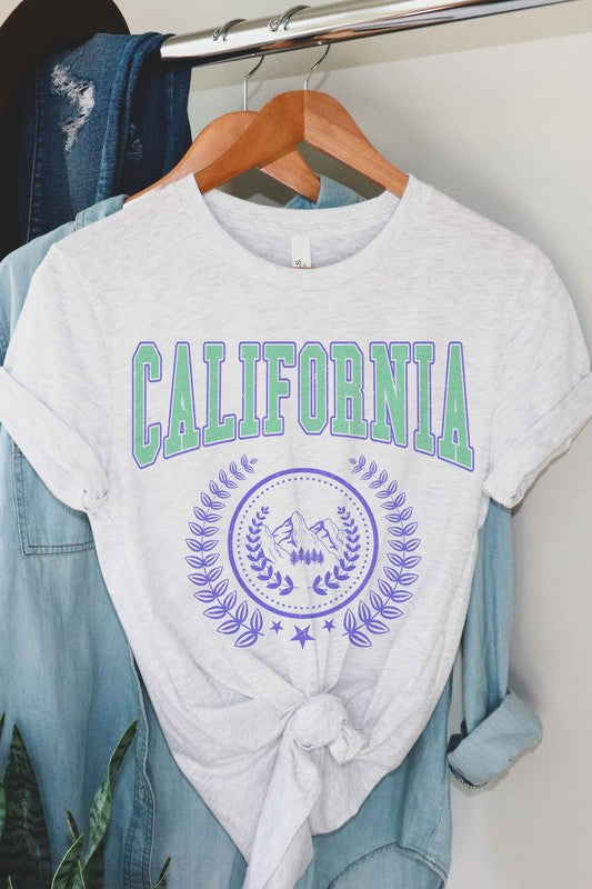 Person wearing a CALIFORNIA GRAPHIC TEE, which features "California" written in green and purple letters along with a mountain and laurel wreath design, paired with white shorts. The T-shirt is unisex and available in plus sizes.