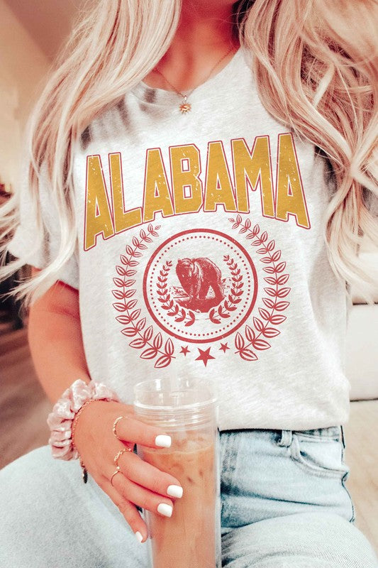 Person wearing an Alabama Graphic Tee made from 100% cotton, featuring a bear emblem, holding a drink in one hand.