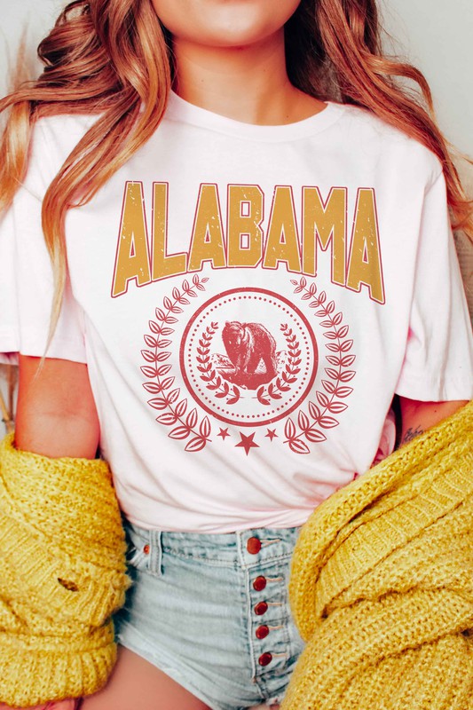 Person wearing an Alabama Graphic Tee made from 100% cotton, featuring a bear emblem, holding a drink in one hand.