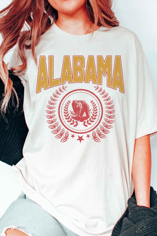 Person wearing an Alabama Graphic Tee made from 100% cotton, featuring a bear emblem, holding a drink in one hand.