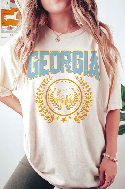 A person is wearing a GEORGIA GRAPHIC TEE in beige, featuring "GEORGIA" in large blue letters above an illustration of a building encircled by laurel wreaths. The unisex tee fits all sizes and the person has long, wavy hair adorned with bracelets. Plus size options are available.