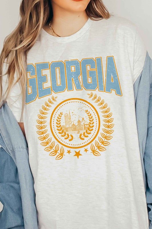 A person is wearing a GEORGIA GRAPHIC TEE in beige, featuring "GEORGIA" in large blue letters above an illustration of a building encircled by laurel wreaths. The unisex tee fits all sizes and the person has long, wavy hair adorned with bracelets. Plus size options are available.