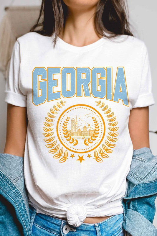 A person is wearing a GEORGIA GRAPHIC TEE in beige, featuring "GEORGIA" in large blue letters above an illustration of a building encircled by laurel wreaths. The unisex tee fits all sizes and the person has long, wavy hair adorned with bracelets. Plus size options are available.