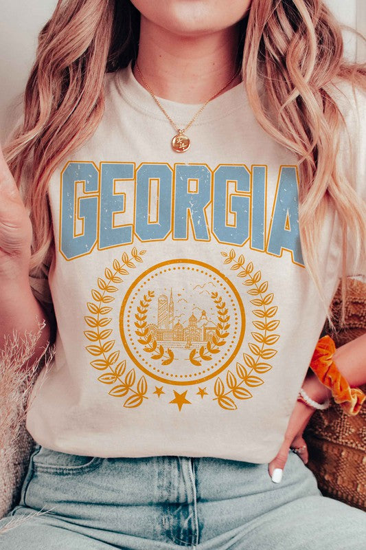 A person is wearing a GEORGIA GRAPHIC TEE in beige, featuring "GEORGIA" in large blue letters above an illustration of a building encircled by laurel wreaths. The unisex tee fits all sizes and the person has long, wavy hair adorned with bracelets. Plus size options are available.