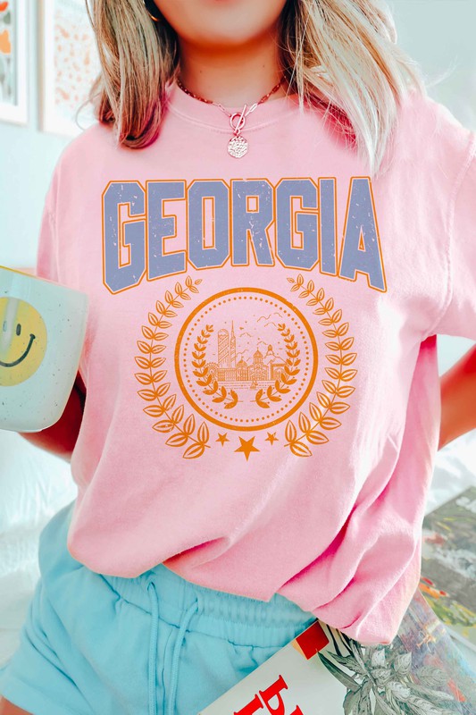 A person is wearing a GEORGIA GRAPHIC TEE in beige, featuring "GEORGIA" in large blue letters above an illustration of a building encircled by laurel wreaths. The unisex tee fits all sizes and the person has long, wavy hair adorned with bracelets. Plus size options are available.