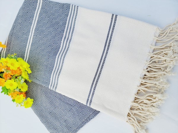 A folded, patterned sand-proof beach towel with a black and white diamond design and a bouquet of purple and pink flowers placed on top. The towel, reminiscent of a Turkish towel, has fringed edges.