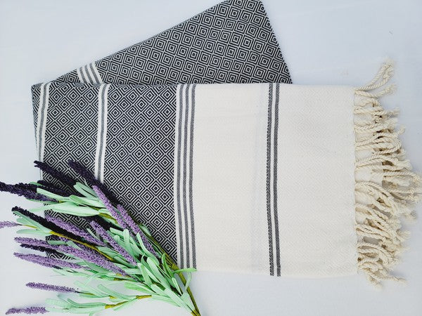 A folded, patterned sand-proof beach towel with a black and white diamond design and a bouquet of purple and pink flowers placed on top. The towel, reminiscent of a Turkish towel, has fringed edges.