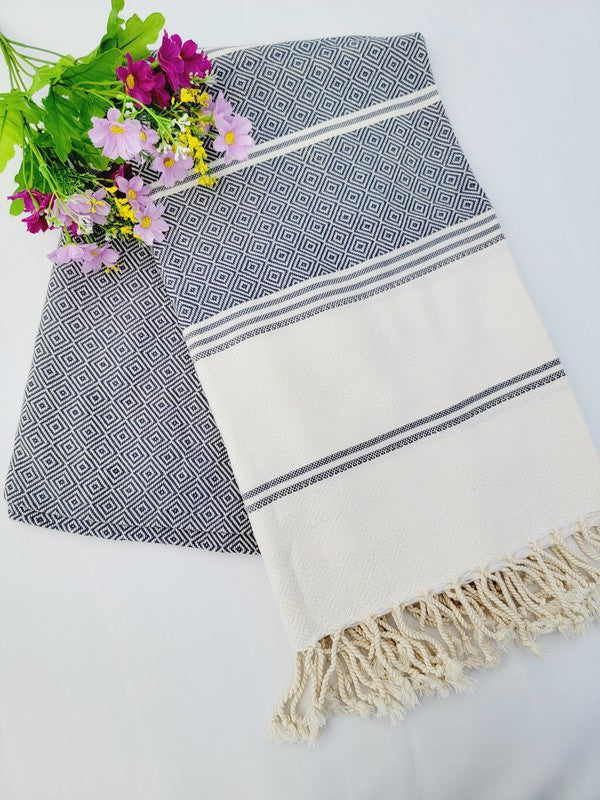 A folded, patterned sand-proof beach towel with a black and white diamond design and a bouquet of purple and pink flowers placed on top. The towel, reminiscent of a Turkish towel, has fringed edges.