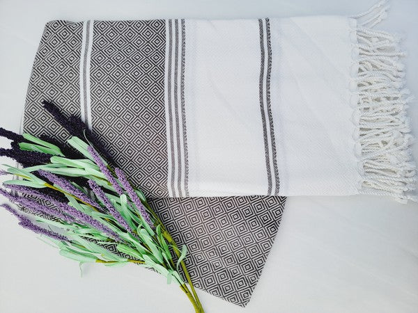 A folded, patterned sand-proof beach towel with a black and white diamond design and a bouquet of purple and pink flowers placed on top. The towel, reminiscent of a Turkish towel, has fringed edges.