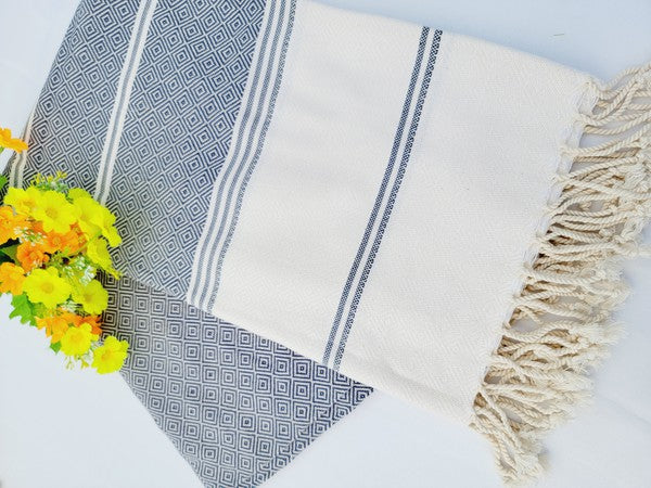 A folded, patterned sand-proof beach towel with a black and white diamond design and a bouquet of purple and pink flowers placed on top. The towel, reminiscent of a Turkish towel, has fringed edges.