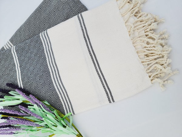 A folded, patterned sand-proof beach towel with a black and white diamond design and a bouquet of purple and pink flowers placed on top. The towel, reminiscent of a Turkish towel, has fringed edges.