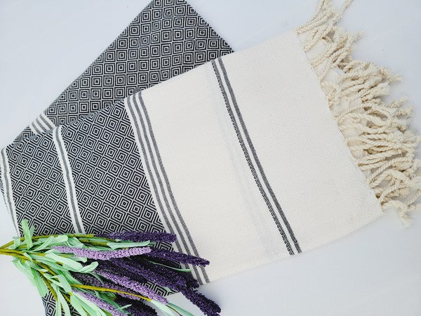 A folded, patterned sand-proof beach towel with a black and white diamond design and a bouquet of purple and pink flowers placed on top. The towel, reminiscent of a Turkish towel, has fringed edges.