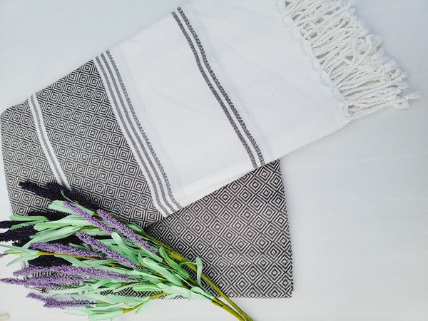 A folded, patterned sand-proof beach towel with a black and white diamond design and a bouquet of purple and pink flowers placed on top. The towel, reminiscent of a Turkish towel, has fringed edges.