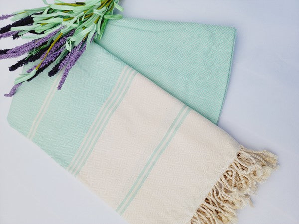 A sand-proof Turkish towel with beige and green stripes and fringed edges lies next to a folded green fabric featuring a geometric pattern, accompanied by a small lavender bouquet.