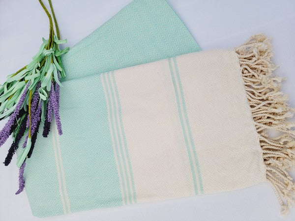 A sand-proof Turkish towel with beige and green stripes and fringed edges lies next to a folded green fabric featuring a geometric pattern, accompanied by a small lavender bouquet.