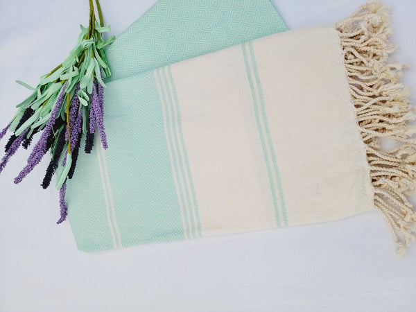 A sand-proof Turkish towel with beige and green stripes and fringed edges lies next to a folded green fabric featuring a geometric pattern, accompanied by a small lavender bouquet.