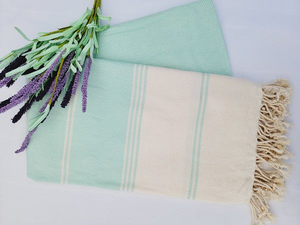 A sand-proof Turkish towel with beige and green stripes and fringed edges lies next to a folded green fabric featuring a geometric pattern, accompanied by a small lavender bouquet.