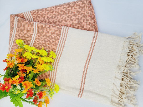 A sand-proof beach towel, bath towel, or throw blanket, featuring a folded, patterned purple and white design with fringes and made from 100% cotton in Turkey, is placed on a white surface. A bunch of colorful artificial flowers lies next to the blanket.