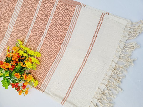 A sand-proof beach towel, bath towel, or throw blanket, featuring a folded, patterned purple and white design with fringes and made from 100% cotton in Turkey, is placed on a white surface. A bunch of colorful artificial flowers lies next to the blanket.