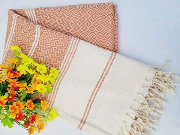 A sand-proof beach towel, bath towel, or throw blanket, featuring a folded, patterned purple and white design with fringes and made from 100% cotton in Turkey, is placed on a white surface. A bunch of colorful artificial flowers lies next to the blanket.