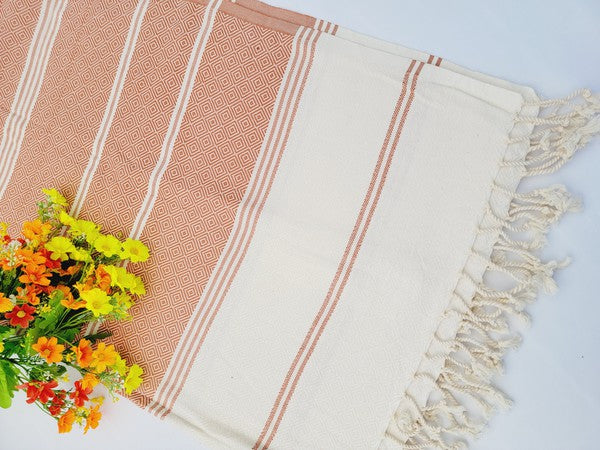 A sand-proof beach towel, bath towel, or throw blanket, featuring a folded, patterned purple and white design with fringes and made from 100% cotton in Turkey, is placed on a white surface. A bunch of colorful artificial flowers lies next to the blanket.