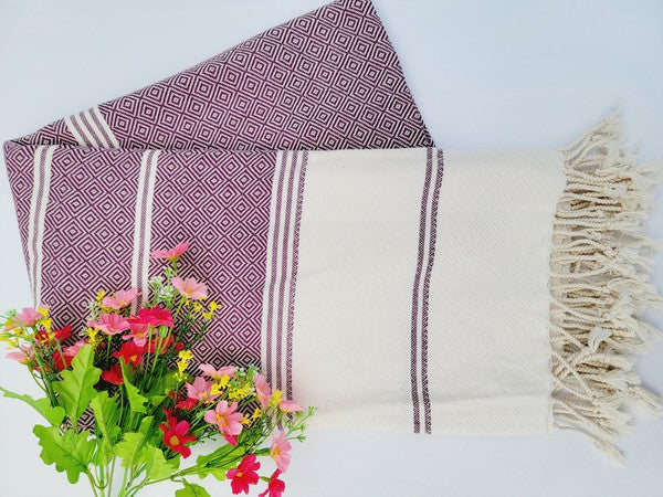 A sand-proof beach towel, bath towel, or throw blanket, featuring a folded, patterned purple and white design with fringes and made from 100% cotton in Turkey, is placed on a white surface. A bunch of colorful artificial flowers lies next to the blanket.