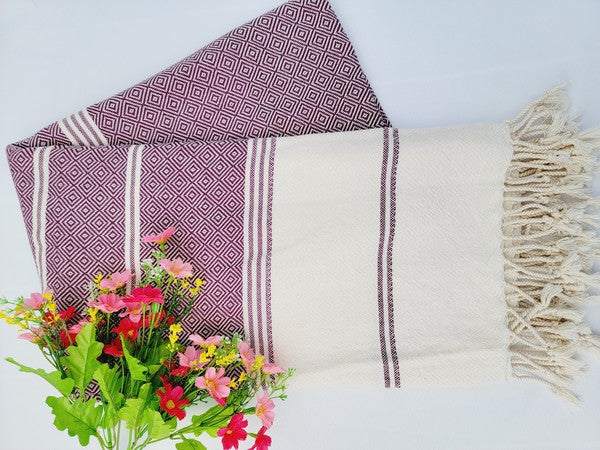 A sand-proof beach towel, bath towel, or throw blanket, featuring a folded, patterned purple and white design with fringes and made from 100% cotton in Turkey, is placed on a white surface. A bunch of colorful artificial flowers lies next to the blanket.