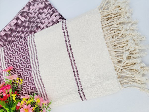 A sand-proof beach towel, bath towel, or throw blanket, featuring a folded, patterned purple and white design with fringes and made from 100% cotton in Turkey, is placed on a white surface. A bunch of colorful artificial flowers lies next to the blanket.