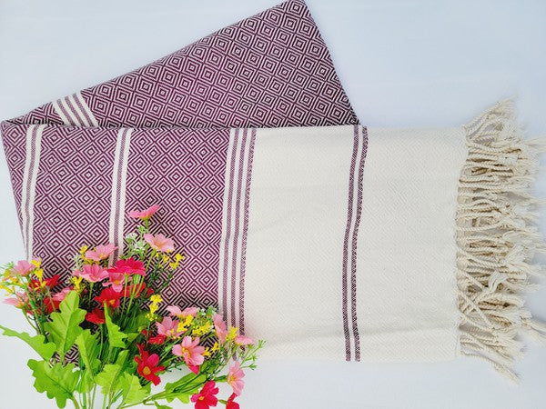 A sand-proof beach towel, bath towel, or throw blanket, featuring a folded, patterned purple and white design with fringes and made from 100% cotton in Turkey, is placed on a white surface. A bunch of colorful artificial flowers lies next to the blanket.