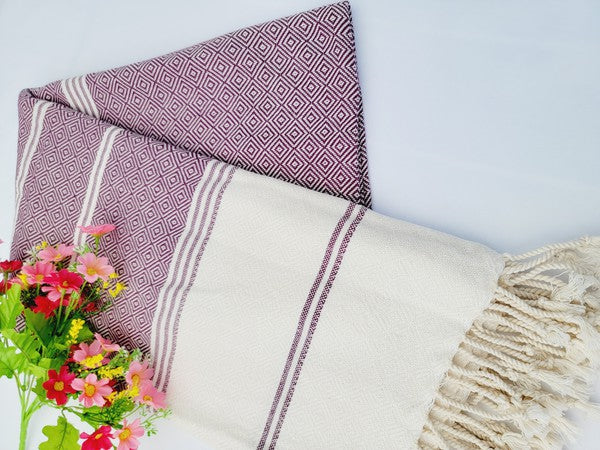 A sand-proof beach towel, bath towel, or throw blanket, featuring a folded, patterned purple and white design with fringes and made from 100% cotton in Turkey, is placed on a white surface. A bunch of colorful artificial flowers lies next to the blanket.