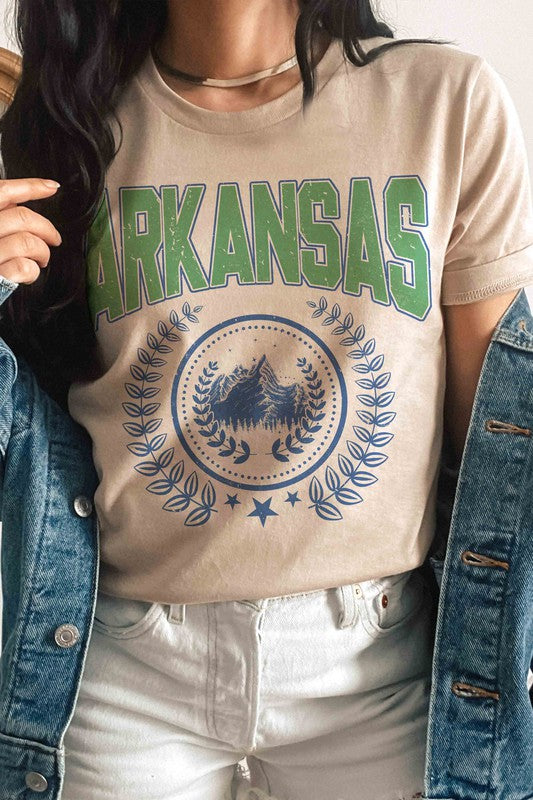 A person wearing a light-colored ARKANSAS GRAPHIC TEE made of 100% cotton and ripped shorts is sitting and holding a white mug.