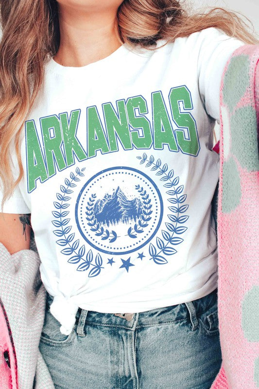 A person wearing a light-colored ARKANSAS GRAPHIC TEE made of 100% cotton and ripped shorts is sitting and holding a white mug.