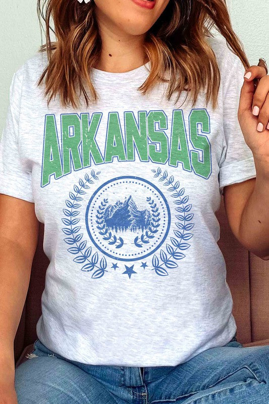 A person wearing a light-colored ARKANSAS GRAPHIC TEE made of 100% cotton and ripped shorts is sitting and holding a white mug.