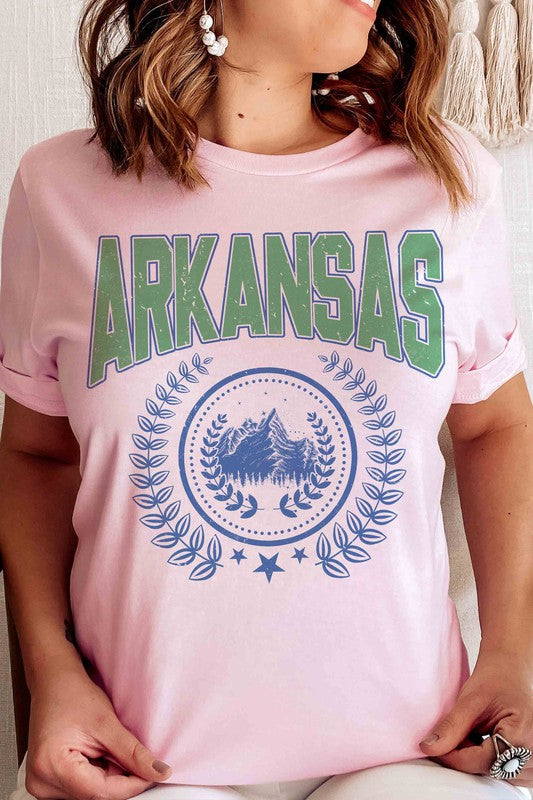 A person wearing a light-colored ARKANSAS GRAPHIC TEE made of 100% cotton and ripped shorts is sitting and holding a white mug.