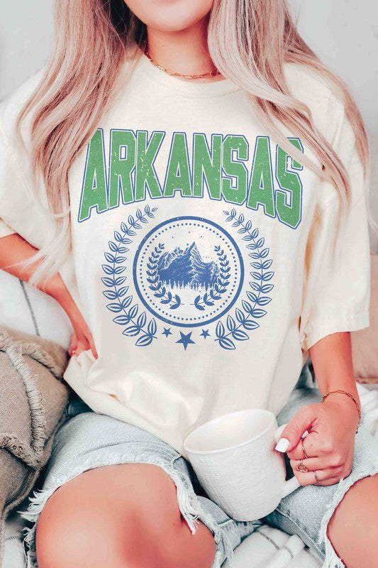 A person wearing a light-colored ARKANSAS GRAPHIC TEE made of 100% cotton and ripped shorts is sitting and holding a white mug.