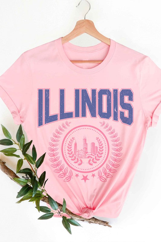 A person is shown wearing an ILLINOIS GRAPHIC TEE, made from 100% cotton, featuring "ILLINOIS" printed in blue above a pink graphic of Chicago's skyline encircled by a decorative laurel wreath; they have wavy hair and are also wearing jeans.