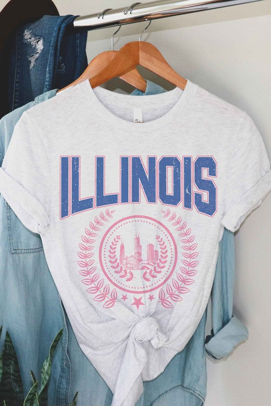 A person is shown wearing an ILLINOIS GRAPHIC TEE, made from 100% cotton, featuring "ILLINOIS" printed in blue above a pink graphic of Chicago's skyline encircled by a decorative laurel wreath; they have wavy hair and are also wearing jeans.