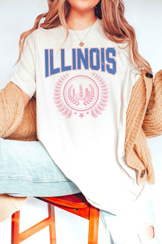 A person is shown wearing an ILLINOIS GRAPHIC TEE, made from 100% cotton, featuring "ILLINOIS" printed in blue above a pink graphic of Chicago's skyline encircled by a decorative laurel wreath; they have wavy hair and are also wearing jeans.